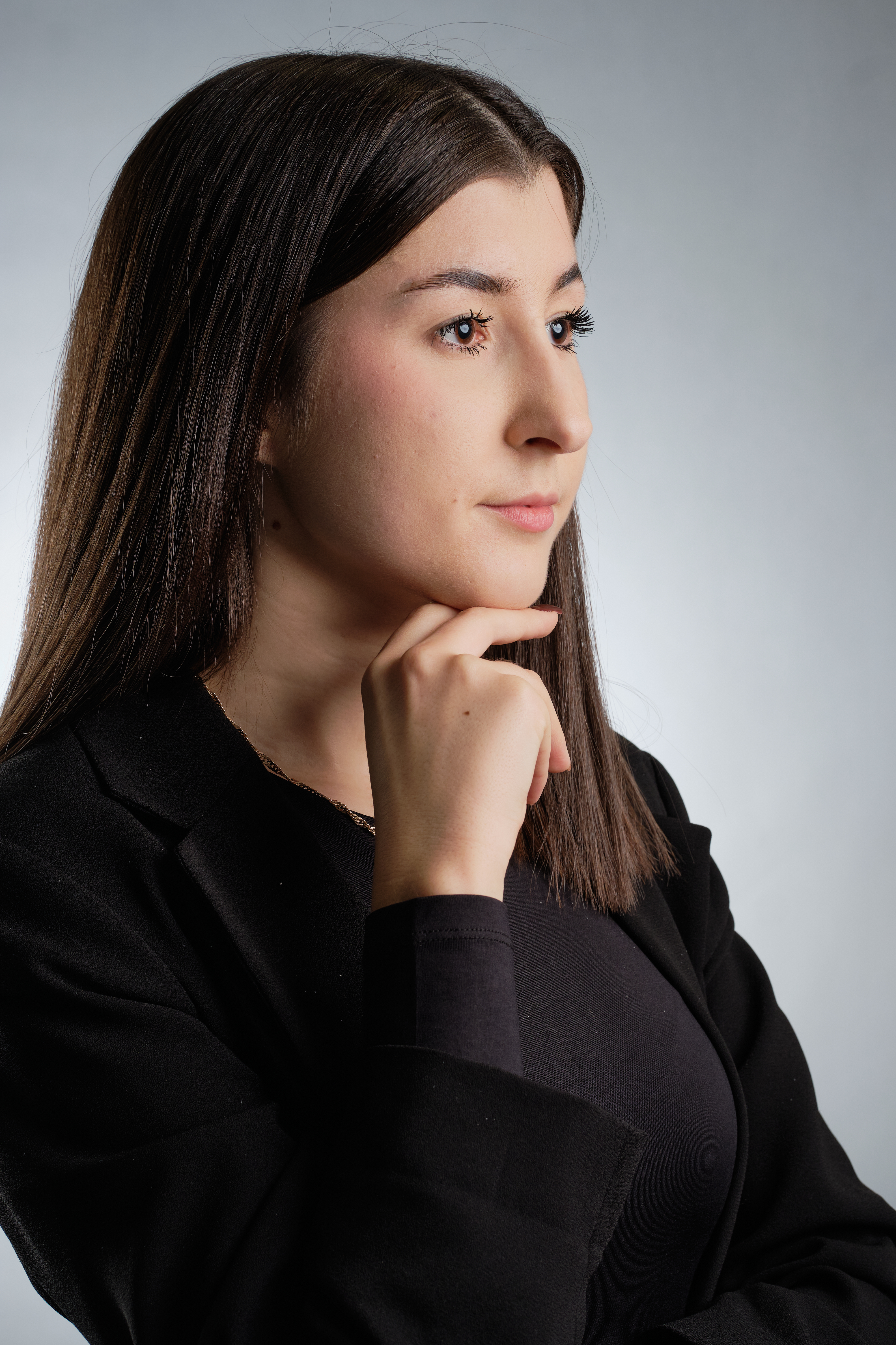 Kobieta w pozie formalnej ubrana w czarną marynarkę, podpiera dłonią podbródek, patrzy w prawo. Portret studyjny