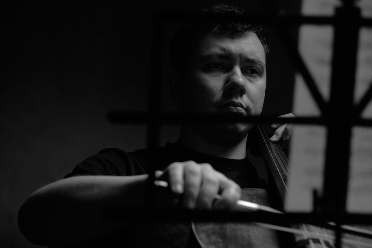 Czarno-biały portret wiolonczelisty widzianego przez konstrukcję pulpitu na nuty, profesjonalna fotografia muzyczna wykonana w Katowicach przez Andrzeja Hanzlika.