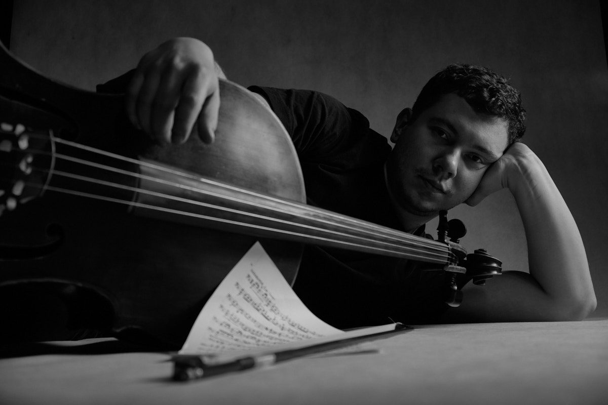 Artystyczna kompozycja czarno-biała: wiolonczelista wsparty na dłoni patrzy w obiektyw, obok niego leżą nuty i smyczek, klimatyczne oświetlenie studyjne, fot. Andrzej Hanzlik. Katowice.os