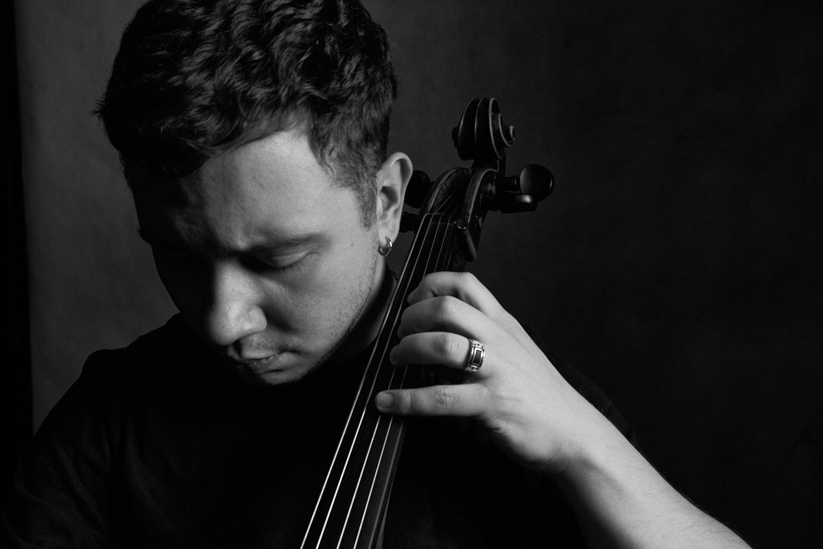 Portret muzyka w czerni i bieli; mężczyzna ze spuszczonym wzrokiem trzyma dłoń na gryfie wiolonczeli, widoczny ozdobny sygnet, oświetlenie typu low-key, fot. Andrzej Hanzlik.